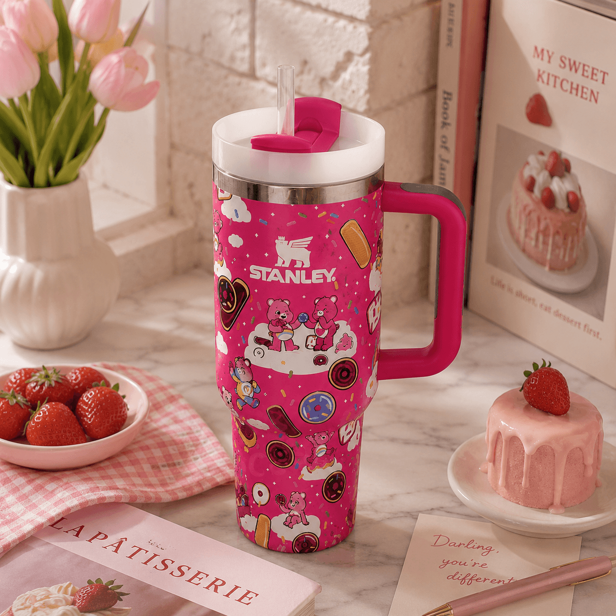 A pink Stanley travel tumbler with a handle and straw, decorated with a whimsical cartoon pattern, styled on a marble countertop with strawberries, a pink cake, and cookbooks in the background.