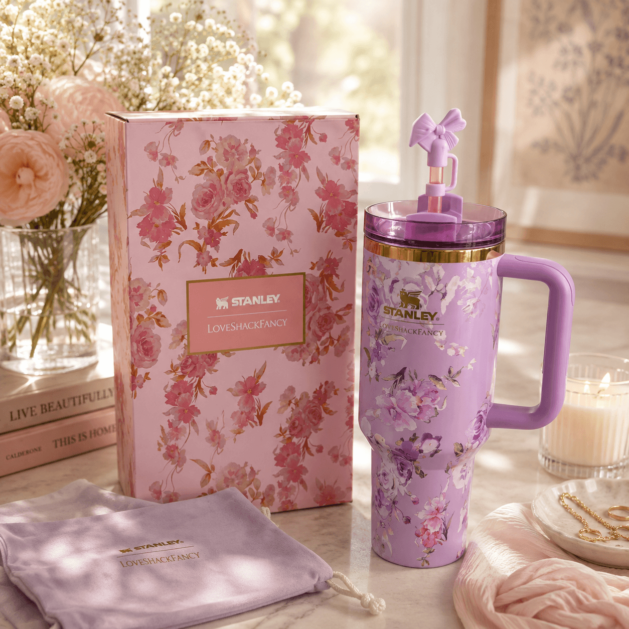A purple floral Stanley tumbler with a handle and lid bow, shown beside its pink floral box and a matching drawstring pouch on a tabletop with flowers and a candle in a softly lit room.