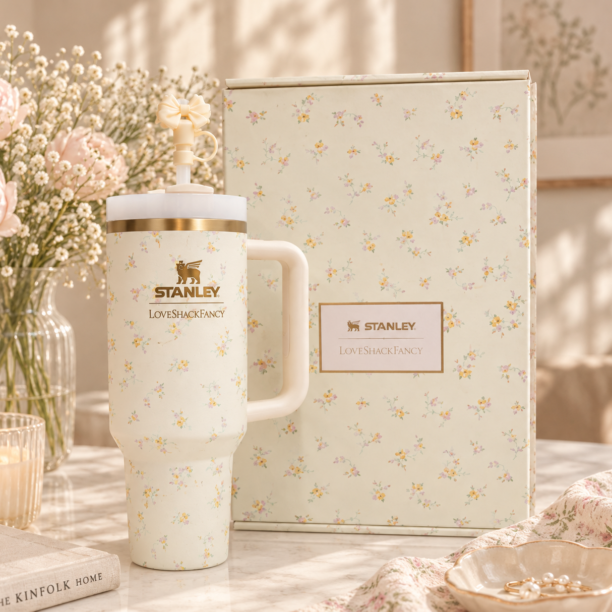A white Stanley tumbler with a delicate yellow and pink floral print, gold band, and gold logo, shown next to its matching floral gift box on a marble table with flowers and a candle in the background.