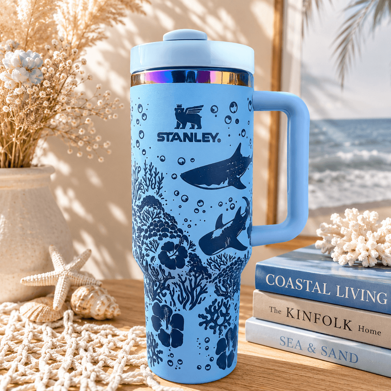 Blue Stanley mug with ocean design on a coastal-themed surface with books and decor.