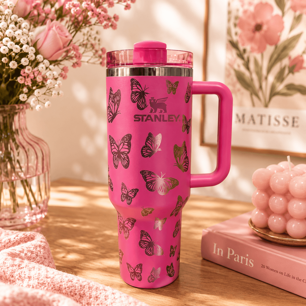 Pink Stanley tumbler with butterfly design on a wooden surface with flowers and books in the background.