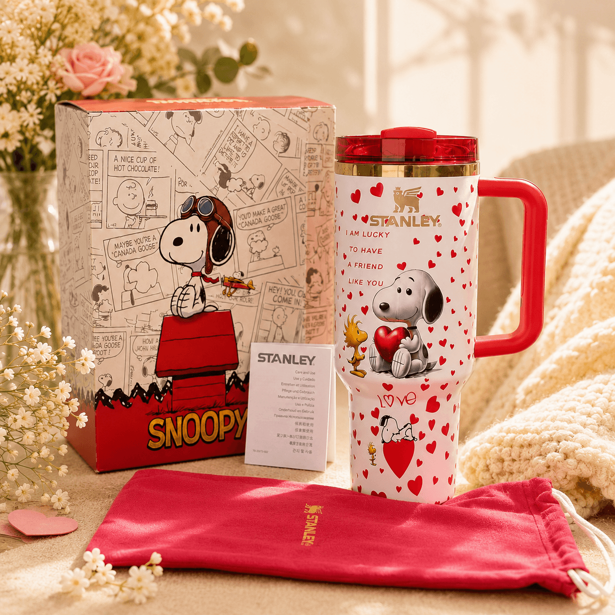 Stanley tumbler with Snoopy design next to a Snoopy-themed box and pink pouch on a decorative background.