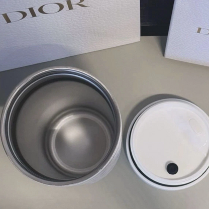 A stainless steel travel tumbler shown open with its white lid placed beside it, with Dior-branded packaging boxes visible in the background.