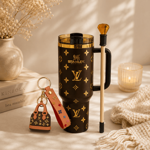 A Stanley travel mug in black with gold monogram pattern, shown with a matching keychain bag charm and a gold-topped metal utensil on a warm neutral tabletop. The scene includes books, a vase with dried flowers, a candle, and a knitted textile in soft natural light.