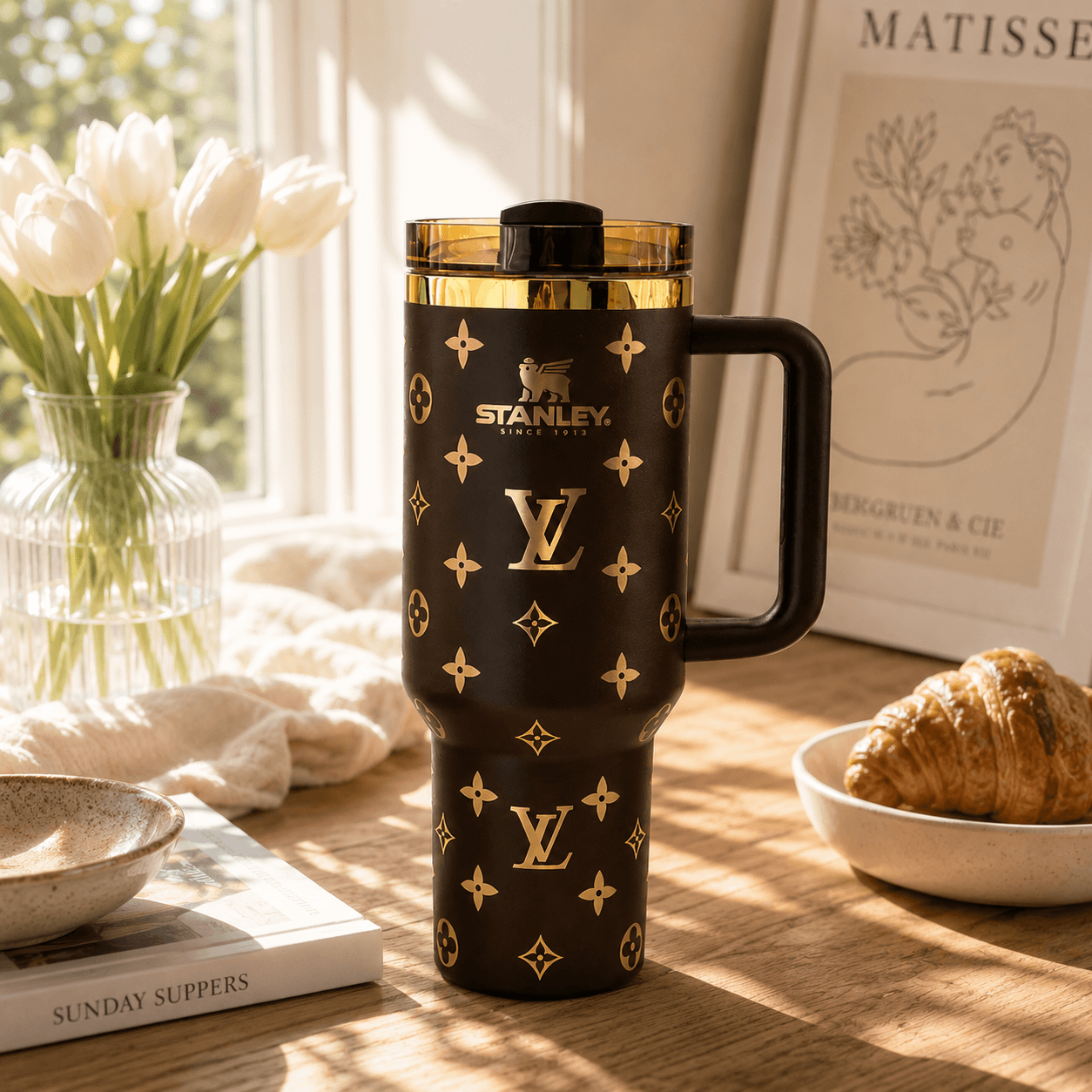 A black Stanley travel mug with gold monogram-style logos and a gold-trimmed lid, shown on a wooden table in a sunlit room with flowers, a book, and a croissant in the background.