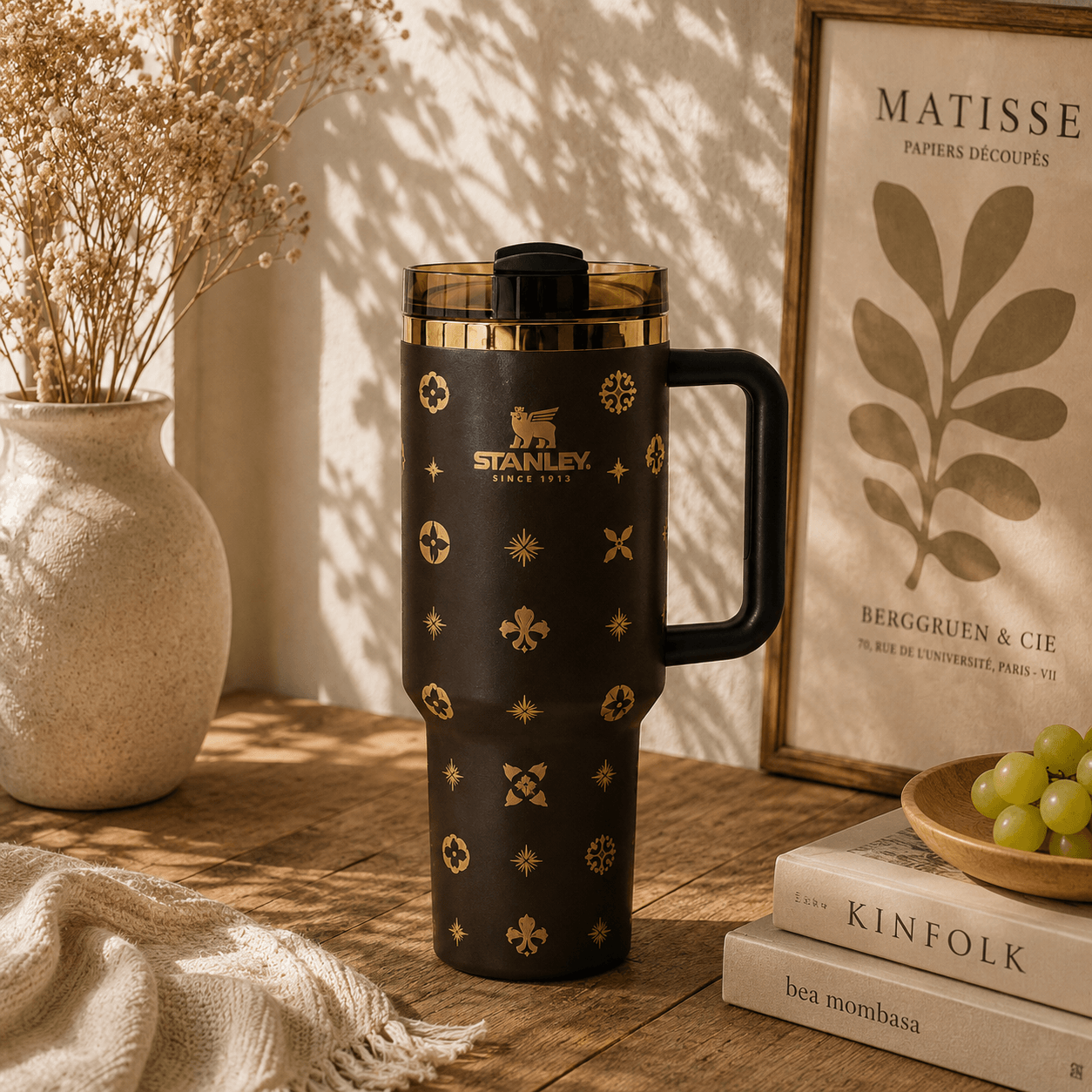 A black Stanley travel mug with gold geometric patterns and a black handle, styled on a wooden table with dried flowers, books, and green grapes in warm sunlight.