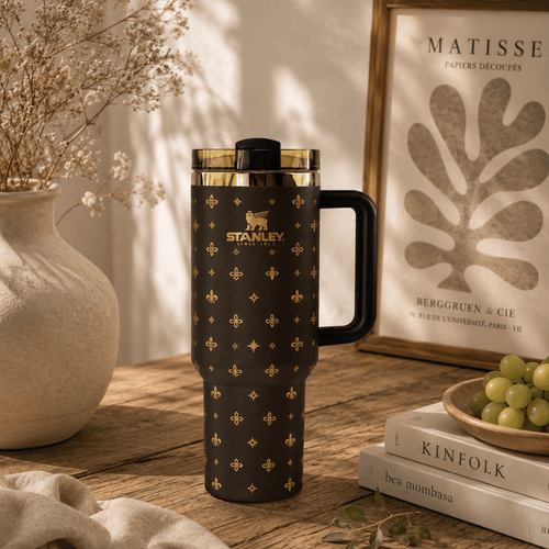 A dark brown Stanley travel tumbler with a gold monogram pattern and black handle, styled on a wooden table with books, grapes, and dried flowers.