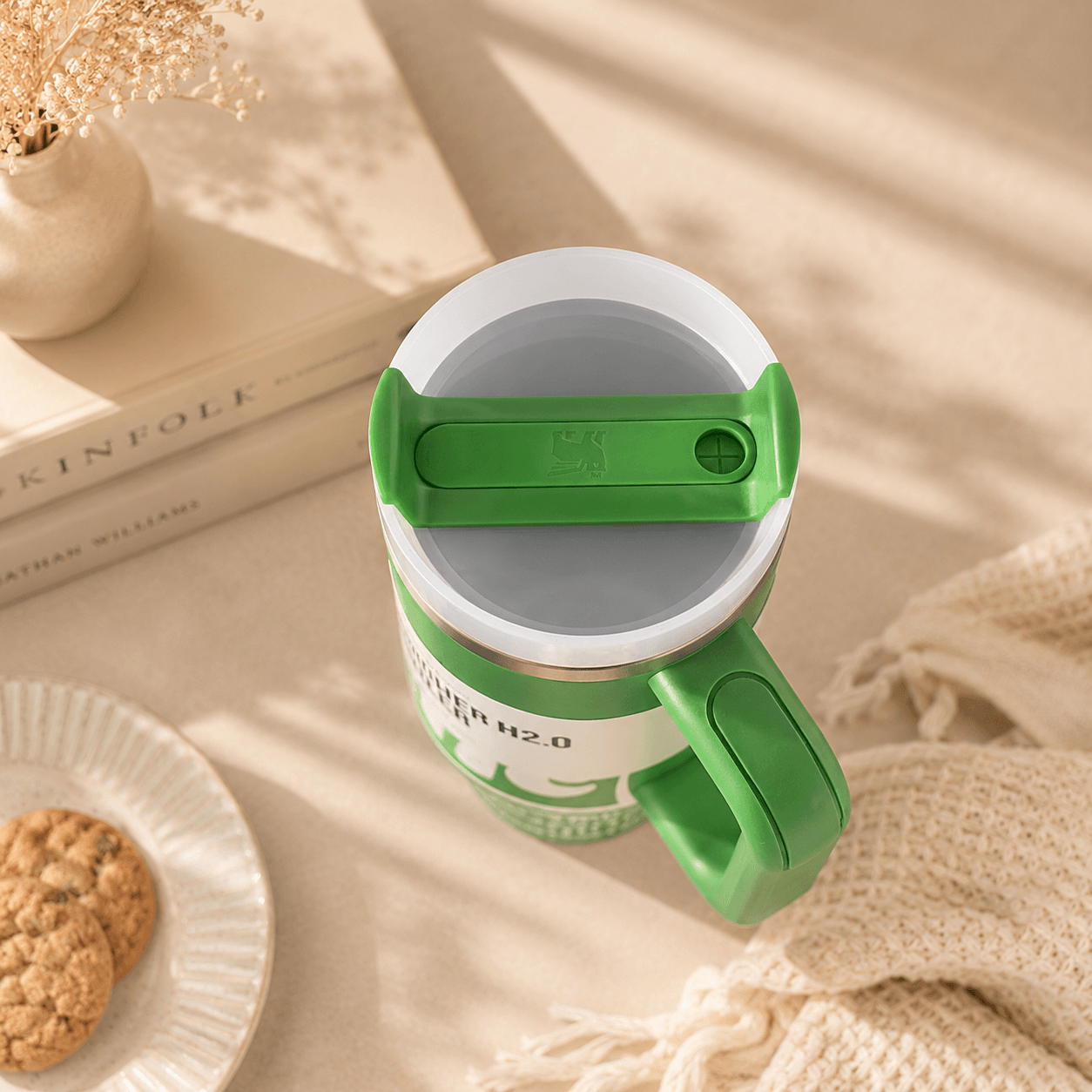 A green and white insulated travel mug with a flip-top lid, styled on a beige surface with a knit blanket, stacked books, and a small plate of cookies.