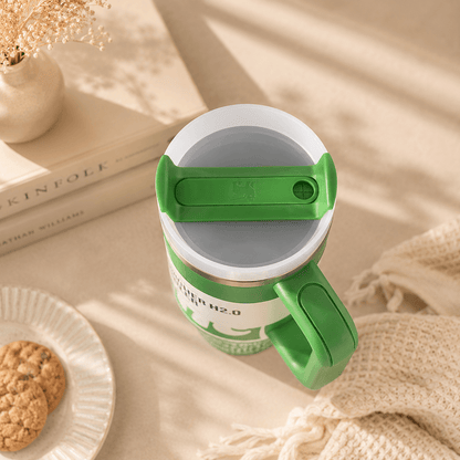 A green and white insulated travel mug with a flip-top lid, styled on a beige surface with a knit blanket, stacked books, and a small plate of cookies.
