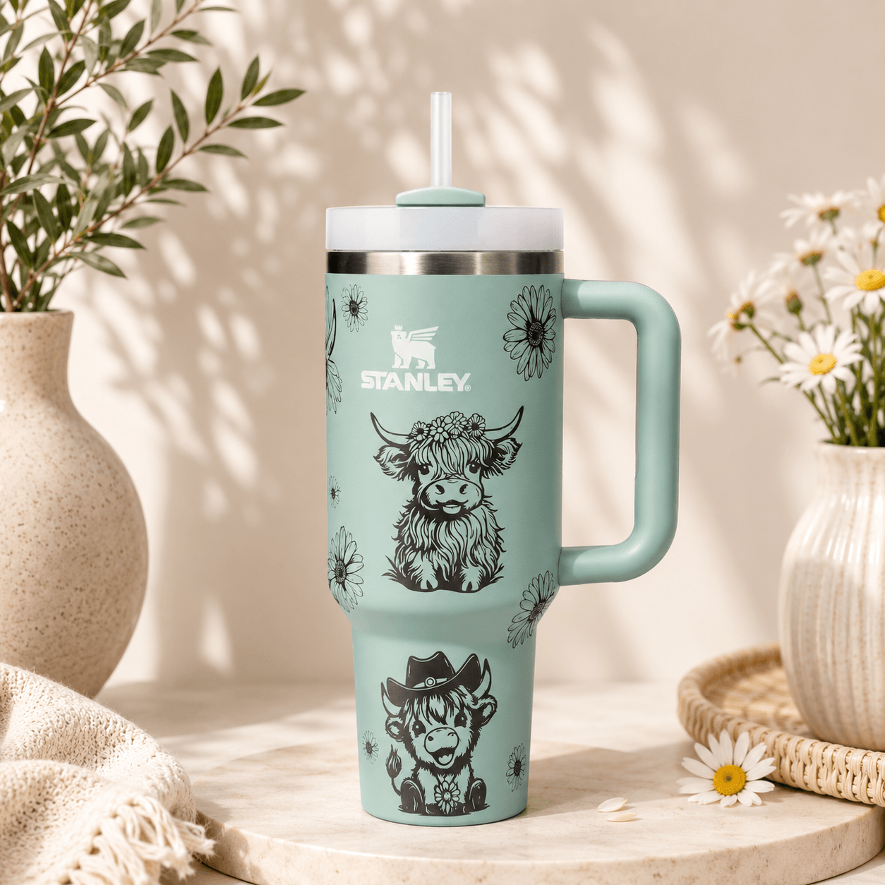 A mint green Stanley tumbler with a handle, lid, and straw, decorated with black cow illustrations and daisy patterns. The tumbler is styled on a stone surface with decorative vases and daisies in the background.