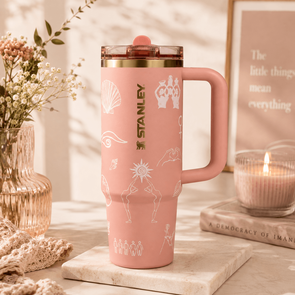 A pink Stanley travel mug with a handle and clear lid, decorated with white line art illustrations, displayed on a marble coaster with a candle, framed print, and flowers in the background.