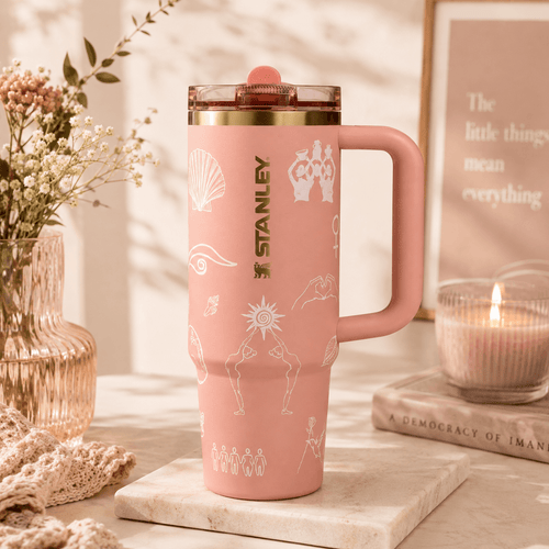 A pink Stanley travel mug with a handle and clear lid, decorated with white line art illustrations, displayed on a marble coaster with a candle, framed print, and flowers in the background.