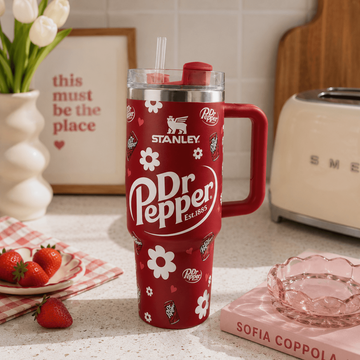 A red Stanley Dr Pepper travel mug with a handle and straw, styled on a kitchen countertop with strawberries, a pink book, and a toaster in the background.