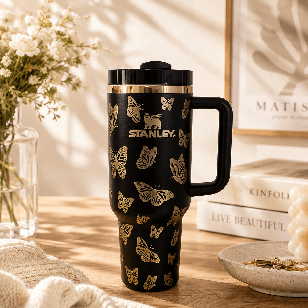 Black Stanley mug with butterfly design on a wooden surface with books and flowers in the background