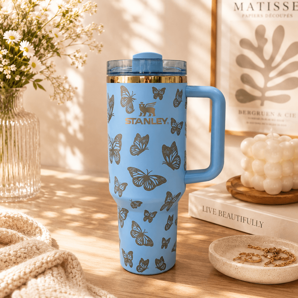 Blue Stanley tumbler with butterfly design on a wooden surface with books and flowers.