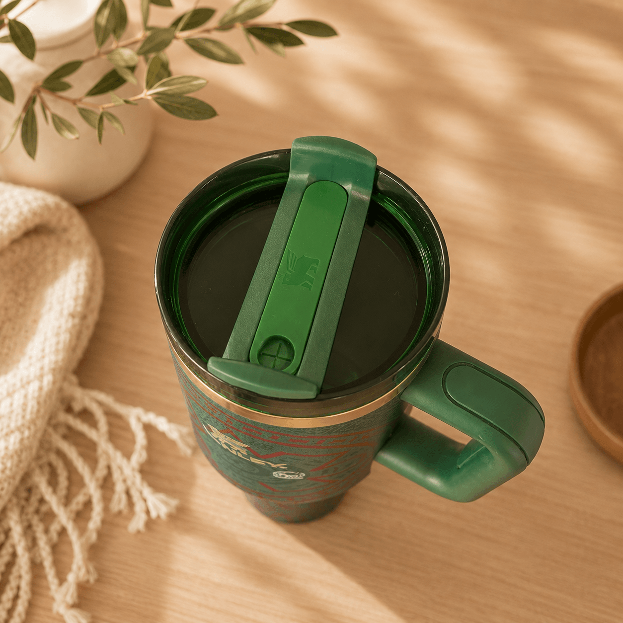Green travel mug with a green lid on a wooden surface with a plant and blanket in the background.
