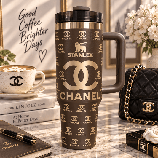 A dark brown Stanley travel mug with gold Chanel logos and text, displayed on a marble table with a Chanel-themed lifestyle setting including a coffee cup, notebook, and handbag.