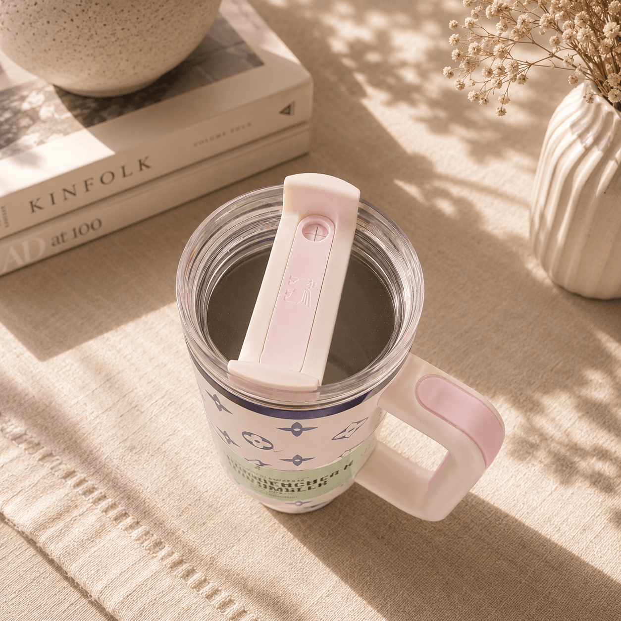 Clear mug with pink handle and white lid on a textured surface with books and decor items.