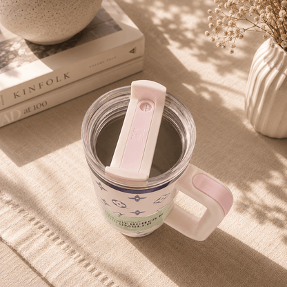 Clear mug with pink handle and white lid on a textured surface with books and decor items.