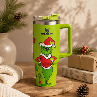 Green Stanley mug with Grinch design on a wooden surface with Christmas decorations.