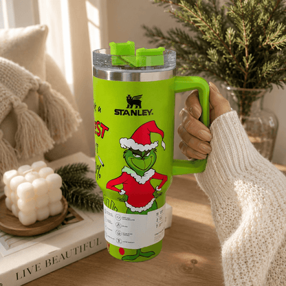 Green Stanley tumbler with Grinch design held by a person in a cozy indoor setting.
