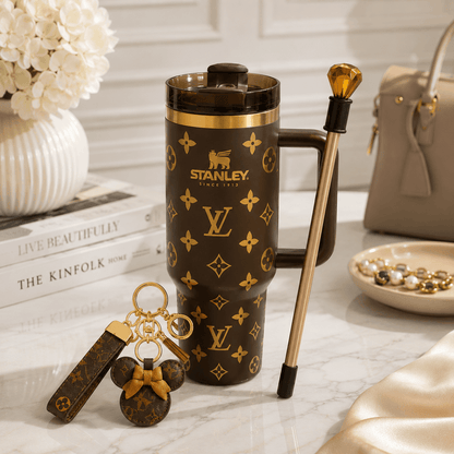 A dark brown Stanley travel mug with gold Louis Vuitton-style monogram print, shown on a marble surface with a matching keychain and a gold-toned pen or utensil beside it. A handbag, books, and flowers are visible in the styled setting.