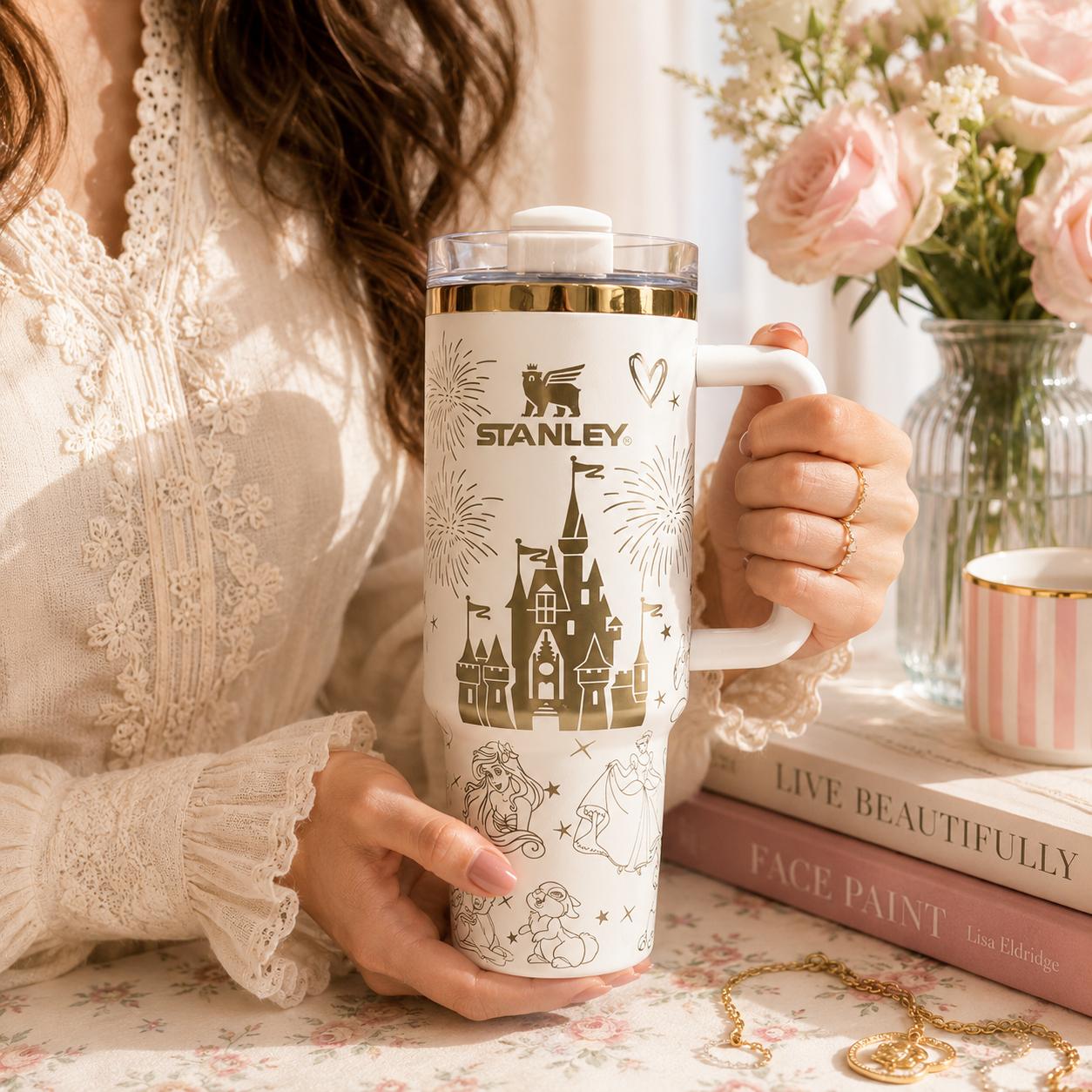 Person holding a Stanley tumbler with a castle design, surrounded by books and flowers.