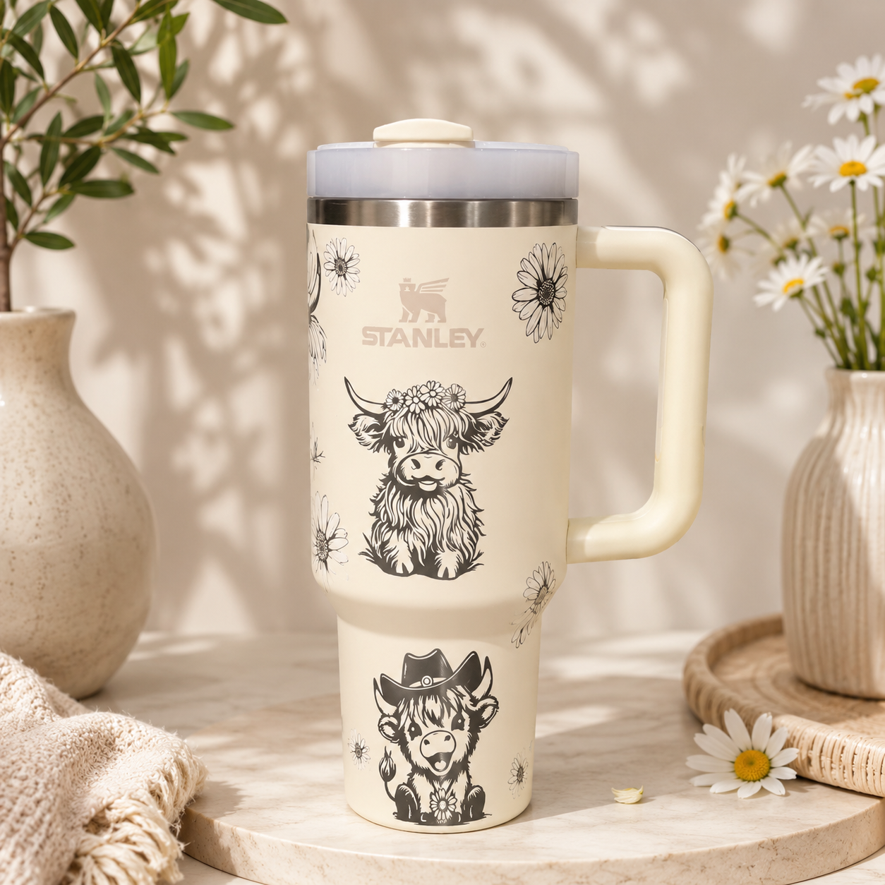 Stanley 40oz insulated tumbler featuring adorable Highland cow illustrations with wildflowers on a cream background, stainless steel construction, ergonomic handle, reusable straw, and double-wall vacuum insulation.