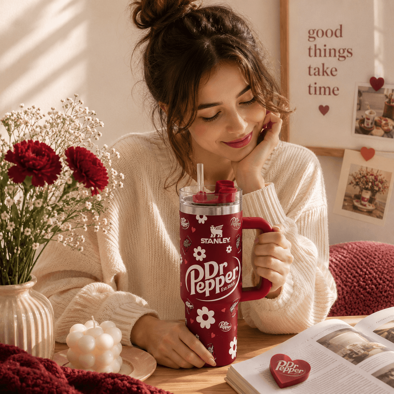 A woman in a cream knit sweater holds a red Stanley Dr Pepper insulated tumbler with a handle and straw, seated at a wooden table with flowers, a candle, and an open book in a warm, cozy room.