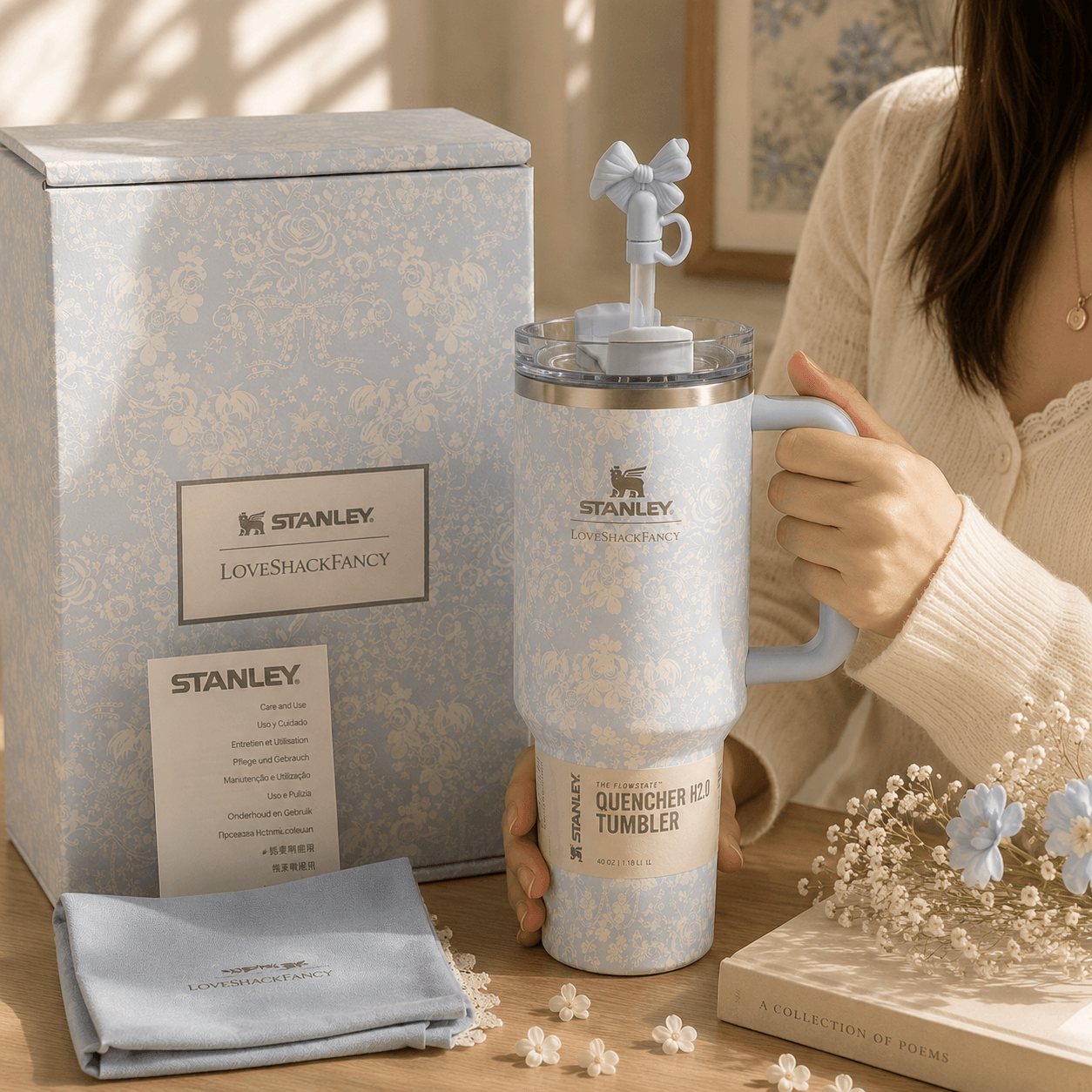 A person holds a light blue and white patterned Stanley Quencher Hot Tumbler with a matching handle and lid, standing beside its branded gift box and a folded cloth on a tabletop.