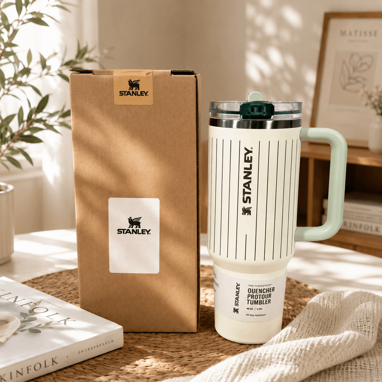 Stanley insulated travel tumbler with a mint green handle and lid, shown next to its brown cardboard packaging box on a woven placemat with a neutral background.