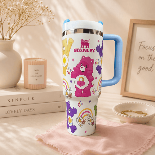 A Stanley insulated travel tumbler with a blue handle, white body, and colorful Care Bear illustrations, styled on a pink cloth with books, a candle, dried flowers, and a framed print in a warm indoor setting.