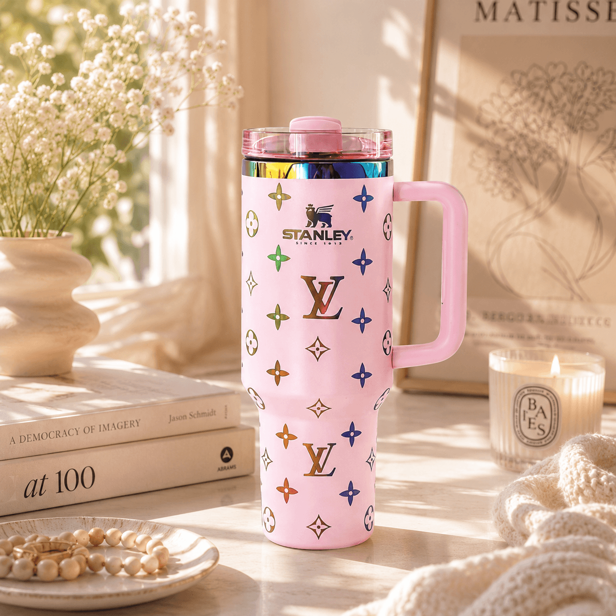 A pink Stanley travel mug with a monogram pattern, featuring a pink handle and lid, styled on a tabletop with books, a candle, and dried flowers.