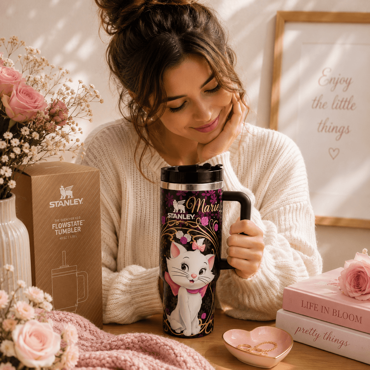 A woman in a cream knit sweater holds a personalized Stanley travel tumbler featuring a white cat illustration and the name 'Marie'. The scene is styled with pink roses, a pink blanket, a pink box of the tumbler, and a framed quote on a warm, sunlit background.