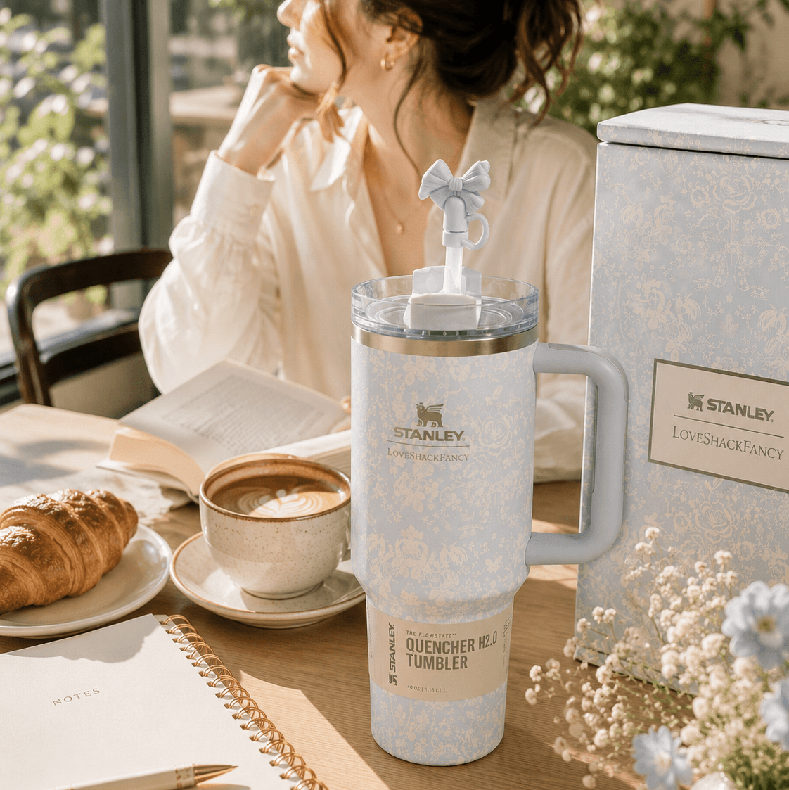 A woman sits at a sunlit table beside a large Stanley tumbler with a handle and straw, next to its branded box. The scene includes a croissant, coffee, notebooks, and small flowers in a warm indoor setting.