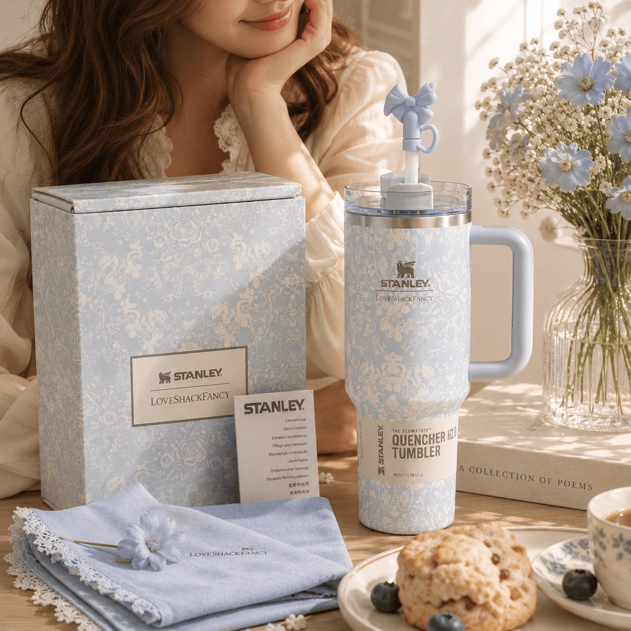 A woman in a cream blouse sits beside a light blue patterned Stanley tumbler with a handle and lid, next to a matching blue gift box and folded napkin on a table. A vase of blue flowers and a book are in the background.