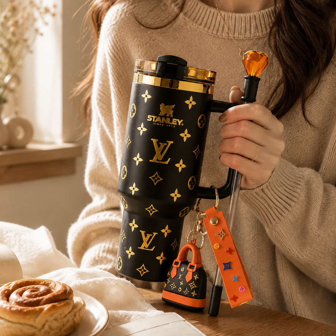 Person holding a Stanley tumbler with decorative pattern, surrounded by a cinnamon roll and glasses on a table.