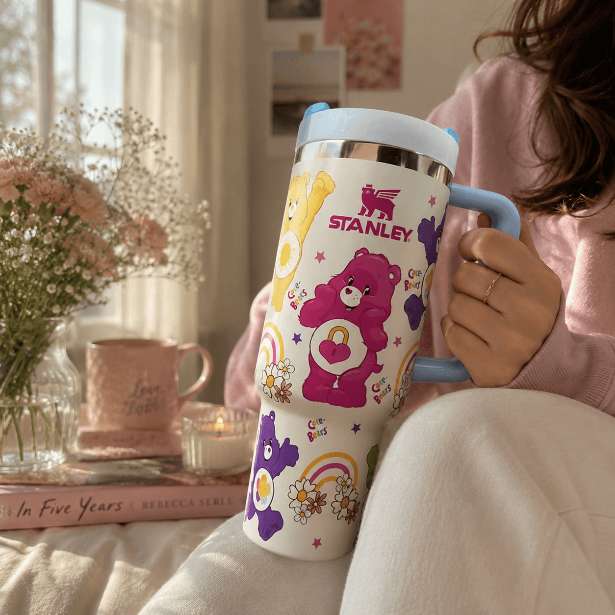 A person in a pink sweater holds a white Stanley insulated tumbler with a light blue lid and handle. The tumbler features colorful Care Bears illustrations, including a pink bear with a heart, a yellow bear, and purple bears, with the Stanley logo printed on the front.