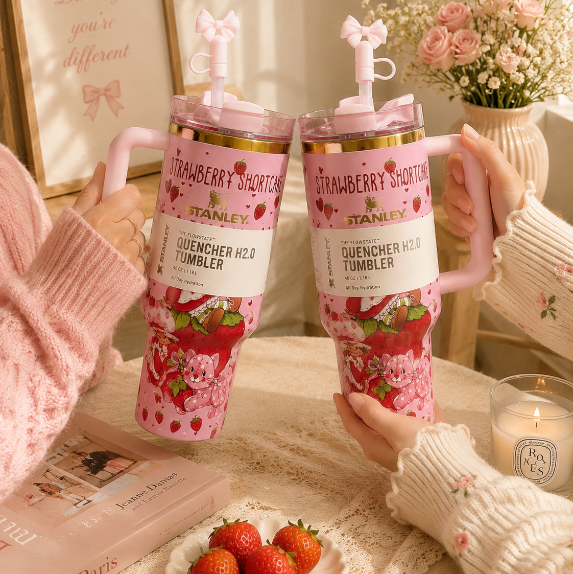 Two pink Strawberry Shortcake-themed tumblers held by people on a table with strawberries and a candle.