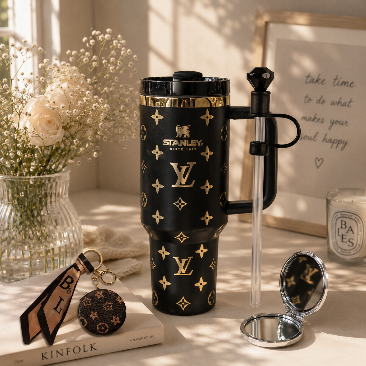 A black Stanley travel tumbler with gold Louis Vuitton-style monogram print, shown on a styled tabletop with a clear straw, compact mirror, keychain, flowers, and framed art in warm sunlight.