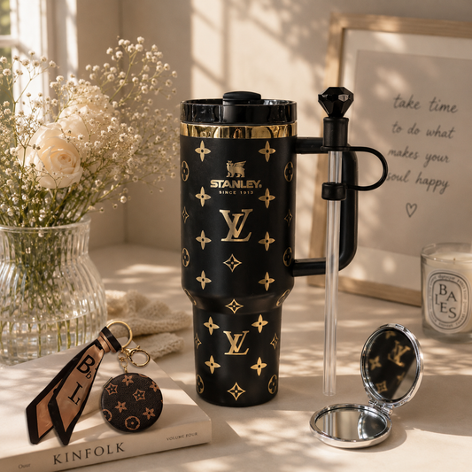A black Stanley travel tumbler with gold Louis Vuitton-style monogram print, shown on a styled tabletop with a clear straw, compact mirror, keychain, flowers, and framed art in warm sunlight.