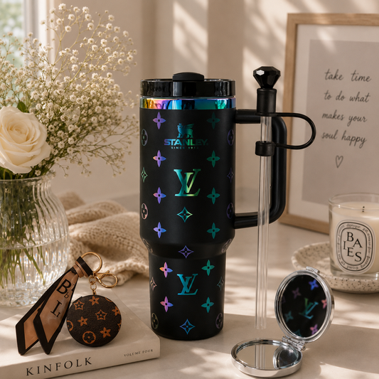 A black insulated travel tumbler with a matching handled straw and a small round mirror, styled on a tabletop with flowers, a framed quote, a candle, and a Louis Vuitton keychain nearby. The tumbler has holographic LV monogram motifs and the visible brand name 'STANLEY SINCE 1904' printed near the top.