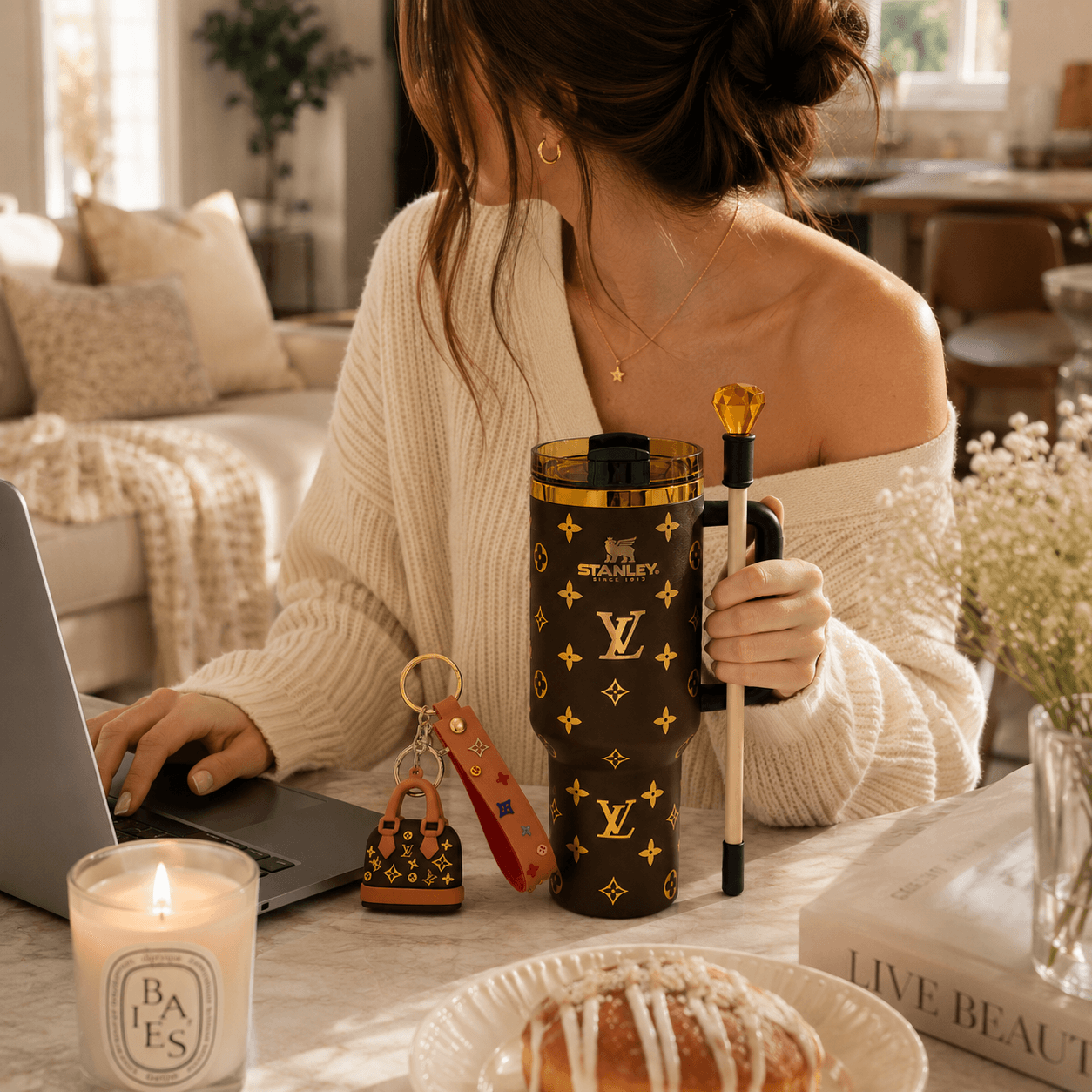 A woman in a cozy off-the-shoulder cream sweater sits at a table in a warm, sunlit room. She holds a dark brown Louis Vuitton Stanley tumbler with gold monogram print and a black-and-white striped pen with a gold top.