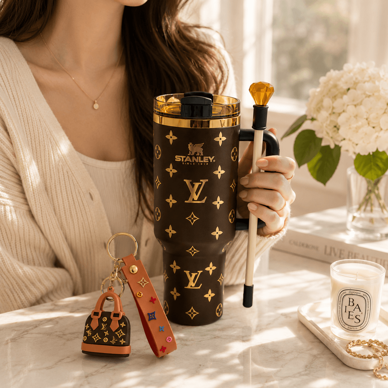 A person in a cream cardigan holds a brown Stanley tumbler with a gold Louis Vuitton-style monogram pattern. A small matching monogram keychain and leather key fob sit on the marble surface beside a lit candle and flowers in the background.