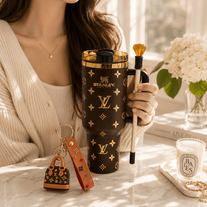 A person in a cream cardigan holds a brown Stanley tumbler with a gold Louis Vuitton-style monogram pattern. A small matching monogram keychain and leather key fob sit on the marble surface beside a lit candle and flowers in the background.