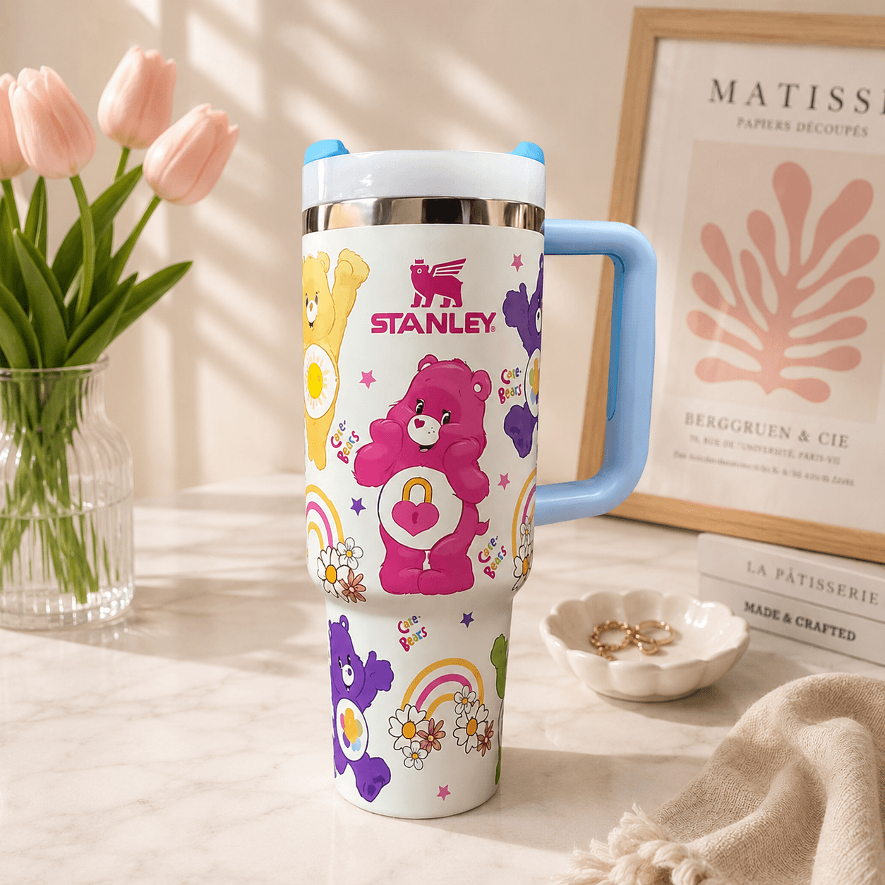 A white insulated tumbler with a light blue handle and lid, decorated with colorful Care Bear graphics and the word 'STANLEY' in red. The tumbler is styled on a marble surface with pink tulips, a framed print, and a small dish in the background.