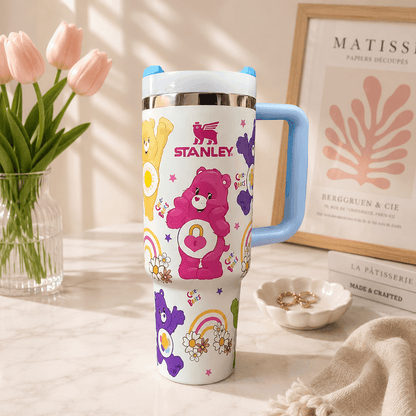 A white insulated tumbler with a light blue handle and lid, decorated with colorful Care Bear graphics and the word 'STANLEY' in red. The tumbler is styled on a marble surface with pink tulips, a framed print, and a small dish in the background.