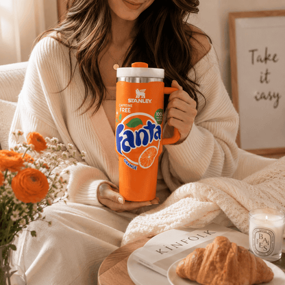 A woman in a cozy cream knit sweater holds an orange Stanley Fanta travel mug in a warm, styled indoor setting with flowers, a croissant, and a candle.