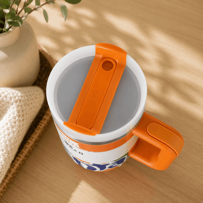 A white insulated mug with a bright orange handle and lid closure, shown on a light wood tabletop with soft shadows from nearby plants.