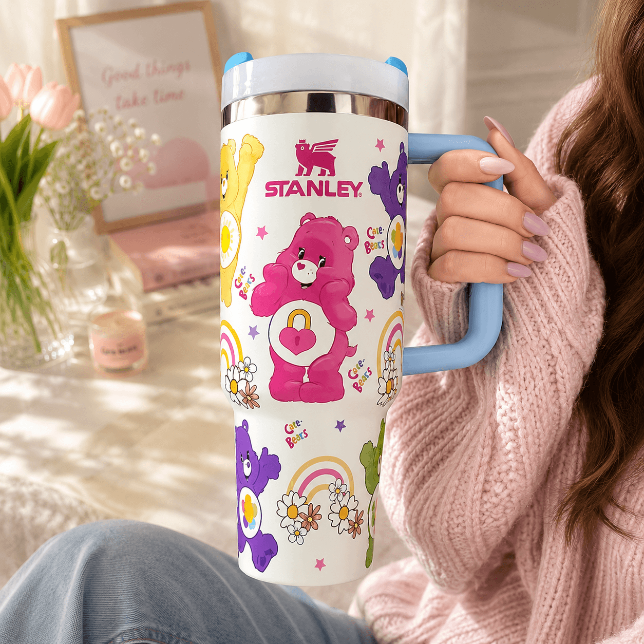 A person in a pink knit sweater holds a Stanley insulated tumbler with a white body, pastel multicolor bear and flower graphics, and a light blue handle. The background is a softly lit indoor setting with flowers and framed decor.